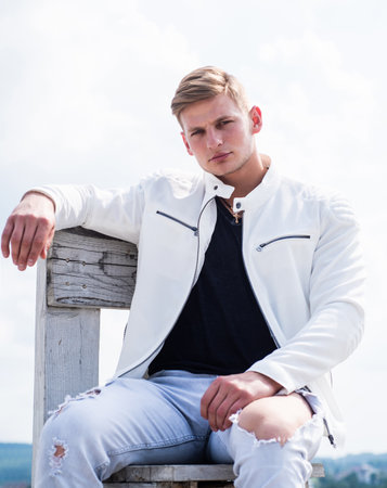 Handsome young man in casual style sit on wooden chair cloudy sky outdoors with pensive look, seriousの写真素材