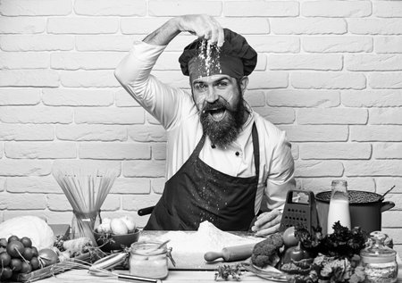 Chef makes dough. Professional cookery concept. Man with beard plays with flour on white brick background. Cook with cheerful face in burgundy uniform sits by table with vegetables and kitchenwarの写真素材