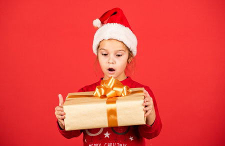 Unbelievable. Amazing surprise. Merry Christmas and happy holidays. Winter holidays. Xmas gift shopping. Gift package. Small adorable girl Santa hat hold gift box. Kid hold present box red backgroundの写真素材