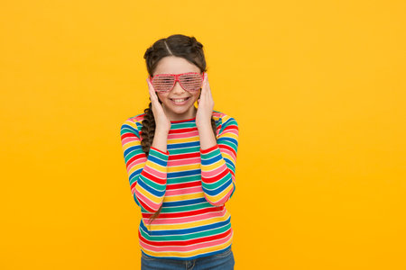 happy kid girl with braided hair wear rainbow color and glamour party glasses on yellow background,...
