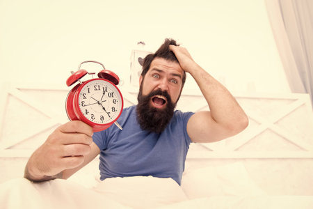 Totally overslept. Get up right now. Stressed man alarm clock. Sleepy man holding alarm clock in bed. Bearded man with alarm clock. Hipster want to sleep. Hate early morning awakening. Daily stressの写真素材