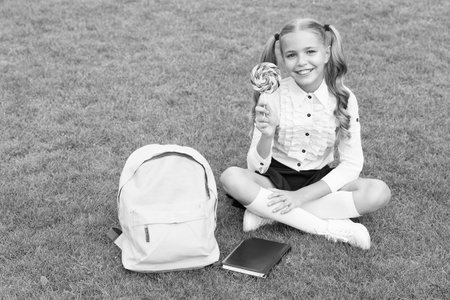 back to school. funny child hold lollipop. concept of education. ordinary learning day. cute girl read book after lesson. happy preschool girl with book in school yardの写真素材