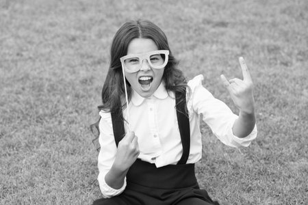 Rock this year. Happy girl hold prop glasses. Beauty look of fashion model. Little child school uniform. Back to school. Small schoolgirl sit on green grass.の写真素材