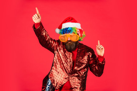 bearded hipster in xmas party glasses. man with beard in santa claus hat. brutal man red background. christmas online shopping. winter holiday party. mature guy celebrate new year. For Santaの写真素材