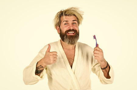 thumb up for success. man in bathrobe brush his teeth. good morning. male health care and personal hygiene. time to relax. brutal bearded hipster isolated on white. feeling happy in bathroomの写真素材