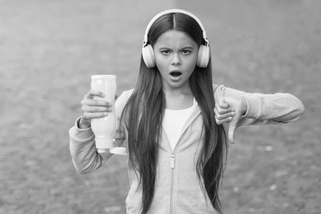 No water no life. Concerned child show thumbs down hand gesture. No water left in bottle. Thirsty girl wear headphones outdoors. Thirst concept. Music and technologyの写真素材