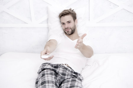 Watch unlimited movies and shows. Subscribe streaming TV services. Guy watching television in bedroom. Online TV concept. Best alternatives to cable TV. Handsome man in pajamas with remote controlの写真素材