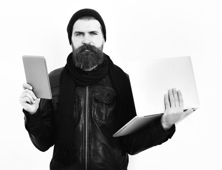 holding tablet and laptop in black leather jacket, hat and scarf isolated on white studio background. Bearded brutal caucasian hipster holding tablet and laptopの写真素材