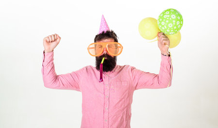 Celebration concept. Man with beard and mustache on busy face blows into party horn, white background. Hipster in giant sunglasses celebrating birthday. Guy in party hat with air balloons celebratesの写真素材