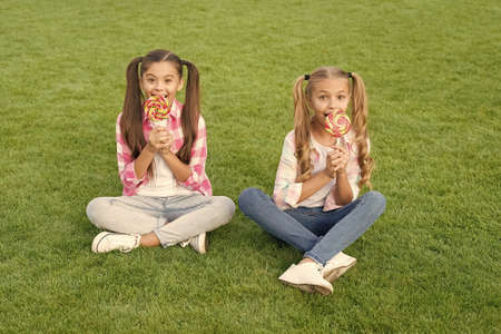 Melt in your mouth dessert. Happy children eat candy dessert on sticks. Little girls sit on green grass. Enjoying sweet dessert. Candy shop. Sugar confectionery. Summer holidays. Dessert time.の写真素材
