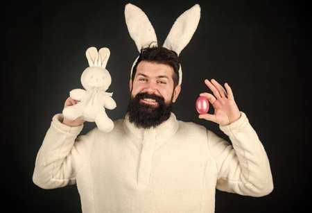 Man wearing rabbit suit. Funny bunny man with easter egg and toy. Easter activities concept. Egg hunt game. Guy bearded hipster cute gentle bunny with long white ears black background. Easter rabbitの写真素材