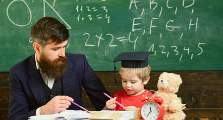 Mathematics lesson concept. Teacher in formal wear and pupil in mortarboard in classroom, chalkboard on background. Enthusiastic kid studying with teacher. Father teaches son mathematicsの写真素材