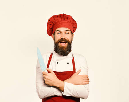 Chef with arms crossed holds sharp blue knife. Cook with happy face in burgundy apron and chef hatの写真素材