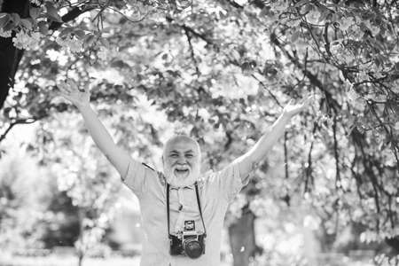Happy grandfather. Capturing beauty life journey. Photographer in blooming garden. Sakura petals falling down. Vintage camera. Retro camera. Senior man hold professional camera. Photography coursesの写真素材