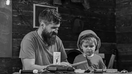 Fatherhood concept. Boy, child busy in protective helmet learning to use hammer with dad. Father, parent with beard teaching little son to use tools, hammering, chalkboard on background.の写真素材
