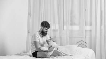 Guy on smiling face playing with teddy bear. Childhood memories concept. Man sits on bed and holds toy, white curtains on background. Macho with beard and mustache relaxing with plush toy after napの写真素材