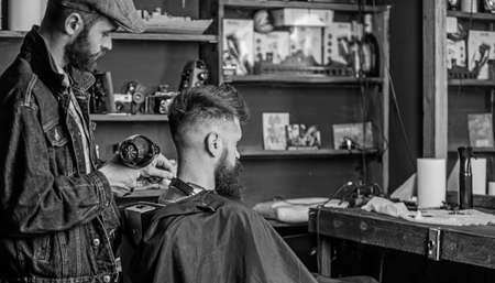 Barbershop concept. Hipster bearded client got hairstyle. Barber with hairdryer works on hairstyle for bearded man, barbershop background. Barber with hairdryer blows off hair out of capeの写真素材