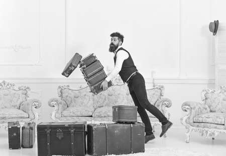 Porter, butler accidentally stumbled, dropping pile of vintage suitcases. Baggage insurance concept. Man with beard and mustache in classic suit delivers luggage, luxury white interior backgroundの写真素材