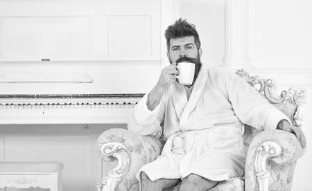 Guy in white bathrobe enjoying morning coffee. Thoughtful man sitting in armchair. Sleepy fellow drinking coffee or tea at homeの写真素材
