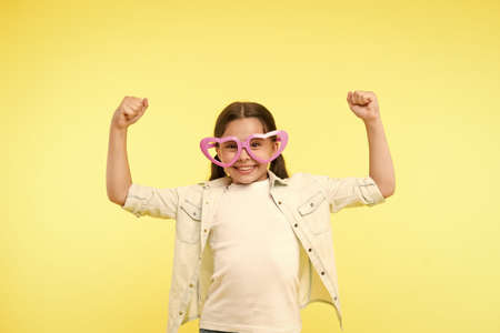 Loves power. Kid girl heart shaped eyeglasses cheerful posing with power gesture. Girl curly hairstyle adorable smiling face. Child charming smile yellow background. Kid happy lovely enjoy childhoodの写真素材