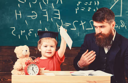 Little boy shows his copybook with paintings. Kid happy studies individually with father, at home. Individual schooling concept. Teacher and pupil in mortarboard, chalkboard on backgroundの写真素材
