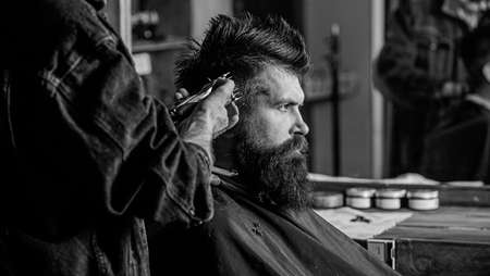 Barber with hair clipper works on hairstyle for bearded man barbershop background. Hipster lifestyle concept. Barber styling hair of brutal bearded client with clipper. Hipster client getting haircutの写真素材