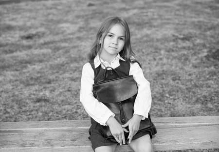 Cute small child with serious look in formal uniform hold school bag sitting on park bench, school...