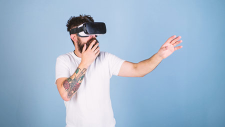 Hipster on shocked face exploring virtual reality with modern gadget. Guy with head mounted display interact in virtual reality. Man with beard in VR glasses, light blue background. VR gadget conceptの写真素材