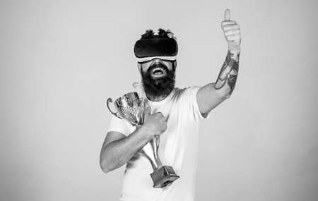 Guy with head mounted display won in virtual game. Man with beard in virtual reality glasses holds golden goblet, light background. Winner concept. Hipster on happy face raising hand with thumb upの写真素材