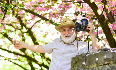 Senior man hold professional camera. Retirement travel. Spring holidays. Travel photo. Photographer in blooming garden. Enjoying free time. Travel and tourism. Capturing beauty. Happy grandfatherの写真素材