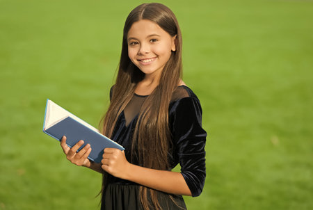 Happy small kid in school uniform smile holding study book green grass sunny summer outdoors, educationの写真素材