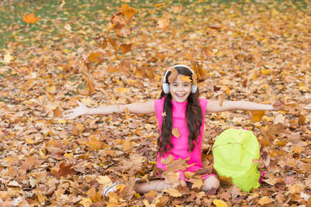 Feeling free and relaxed. child with school backpack in fall forest. concept of knowledge day. kid in autumn park. happy kid throw up autumn leaves. small girl wear headphones. back to schoolの写真素材