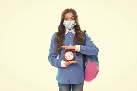 Surprised school child wear protective mask to prevent COVID-19 spread holding alarm clock, deadline