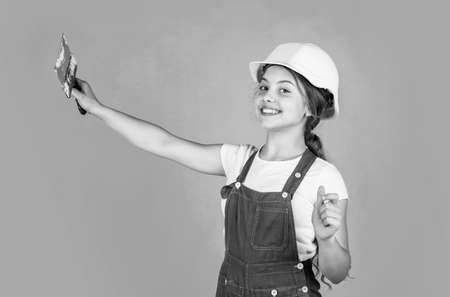 confident kid use spatula while repairing. concept of building and renovation. happy girl use spatula tool. teen builder wear safety helmet. young worker in protective hard hatの写真素材