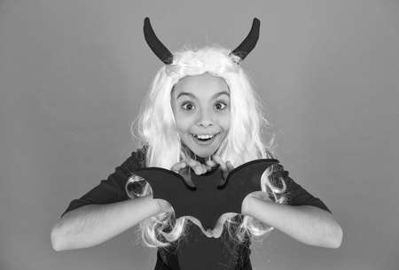 funny kid in witch costume hat for Halloween with devil hors and wig holding bat on orange...