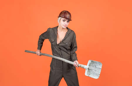 woman laborer in protective helmet and boilersuit hold shovel on orange background, workers dayの写真素材