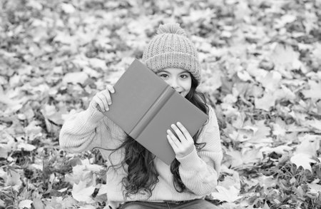 Happy little kid look behind school book sitting on fall leaves outdoors, educationの写真素材