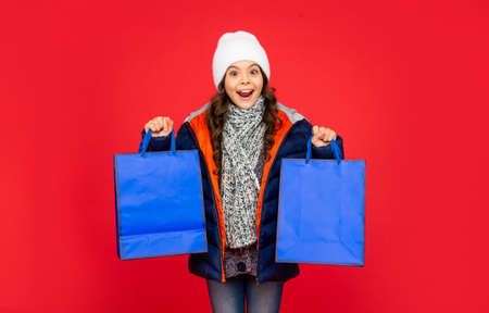 surprised kid in puffer jacket and hat. teen girl after shopping on red backgroundの写真素材