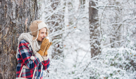 woman winter drink. Girl enjoy snow fall. woman in mittens and hat drinking tea in snowy forest. outdoors on frosty day. travel and hiking. girl hiker drink cup of hot tea. warm yourself upの写真素材