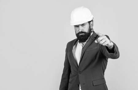 serious man with beard wear hard hat pointing finger. engineering. architecture and building. male builder wearing formal suit and helmet for protection. working on construction project. copy spaceの写真素材