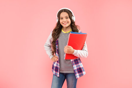 happy child listen music in headphones hold workbook on pink background, education