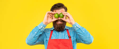 Happy man in apron holding fresh limes on eyes yellow background, fruit sellerの写真素材