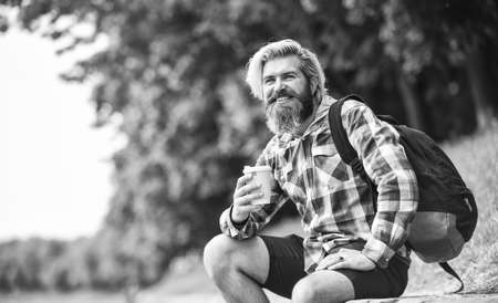 Drinking hot coffee. Drink tea or coffee. Man with cup outdoors. Enjoying nature at riverside. Guy with backpack relaxing. Inspired handsome hipster. Inspiration. Man outdoors with cup of coffeeの写真素材