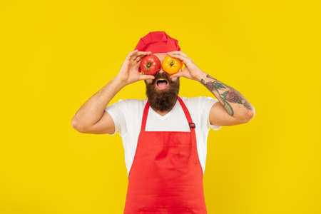 Happy guy in cooking apron and toque holding tomatoes on eyes yellow background, cook