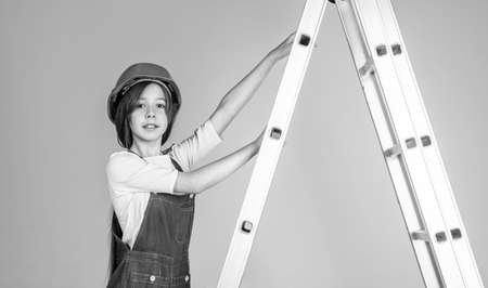 child on ladder wear hard hat. kid builder on construction site. worker engineer. architect in workshop. renovation and repair. teen girl in helmet and boilersuit on stepladderの写真素材