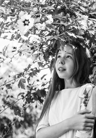 kid spring fashion style. happy childhood. cheerful pretty child at blooming tree outdoor. vacation time. summer fashion and beauty. beautiful teen girl with hibiscus bush flowerの写真素材