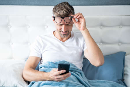 mature man in bed chatting on phone wearing eyeglasses. eyecare