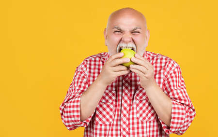 mature man in checkered shirt eating apple on yellow backgroundの写真素材