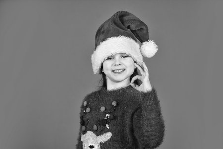 Can not hold back emotions. Counting days till christmas. Christmas party. Christmas holiday invitation. Adorable girl wear santa claus hat red background. Emotional face expression. Happy childhoodの写真素材