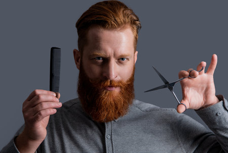 portrait of man has beard and hairstyle in studio. man hold barber scissors and comb.の写真素材